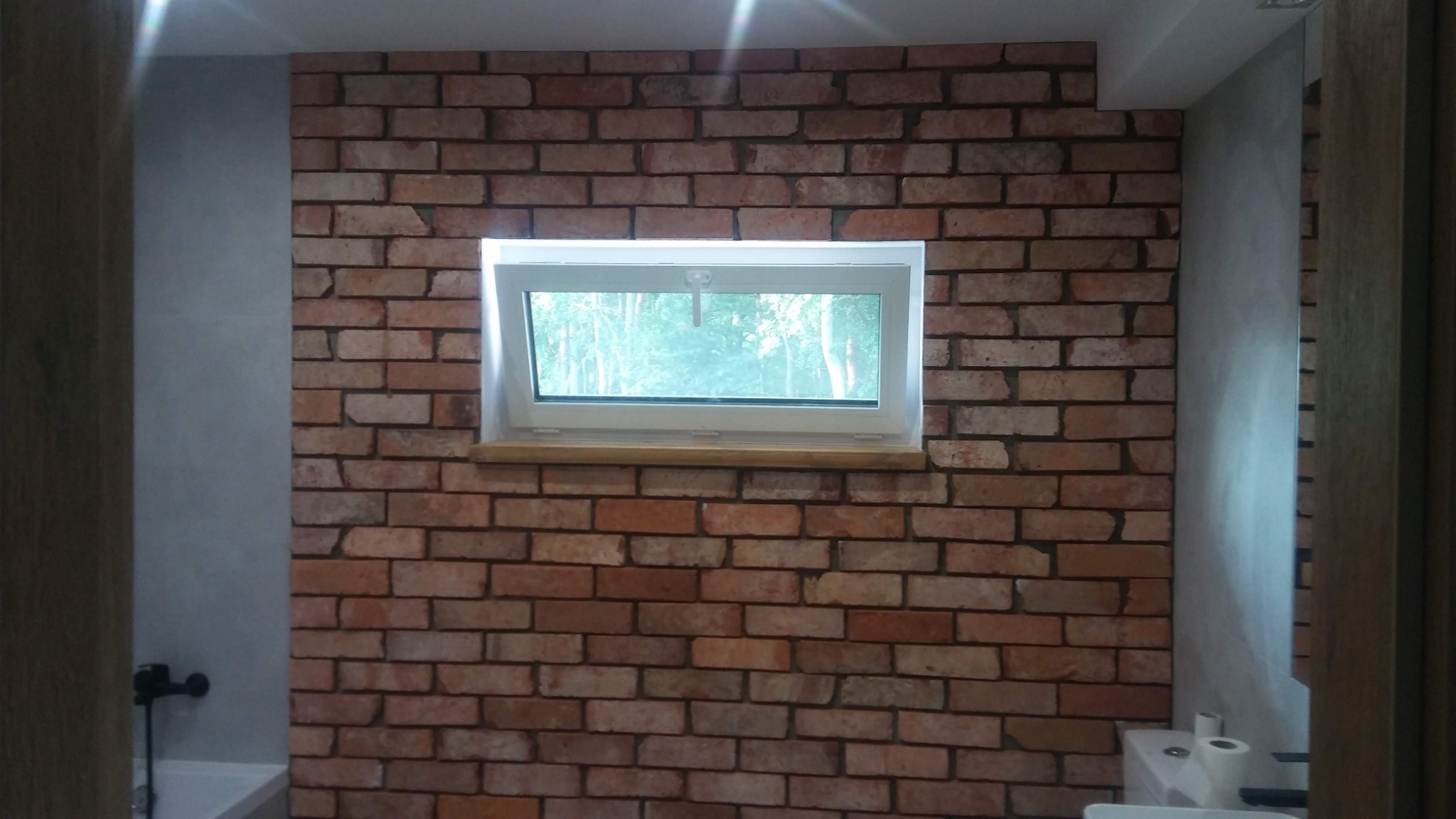 Ściana w łazience wyłożona cegłą klinkierową z oknem uchylnym, widok na zieleń za oknem, obok ściana wykończona gładzią.