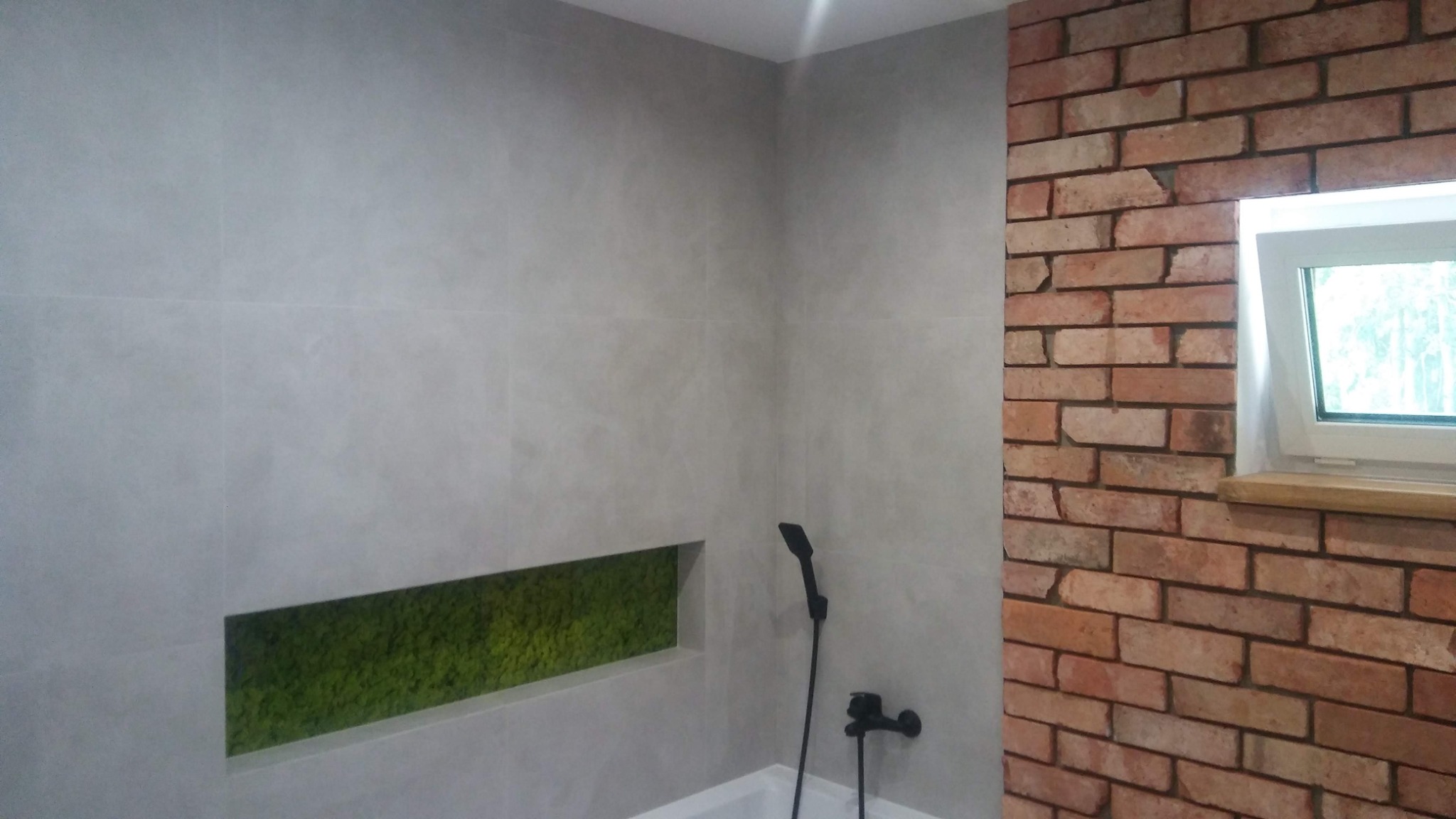 Nowoczesna łazienka z szarymi płytkami imitującymi beton, wnęką z mchem dekoracyjnym i ścianą z cegły przy oknie.