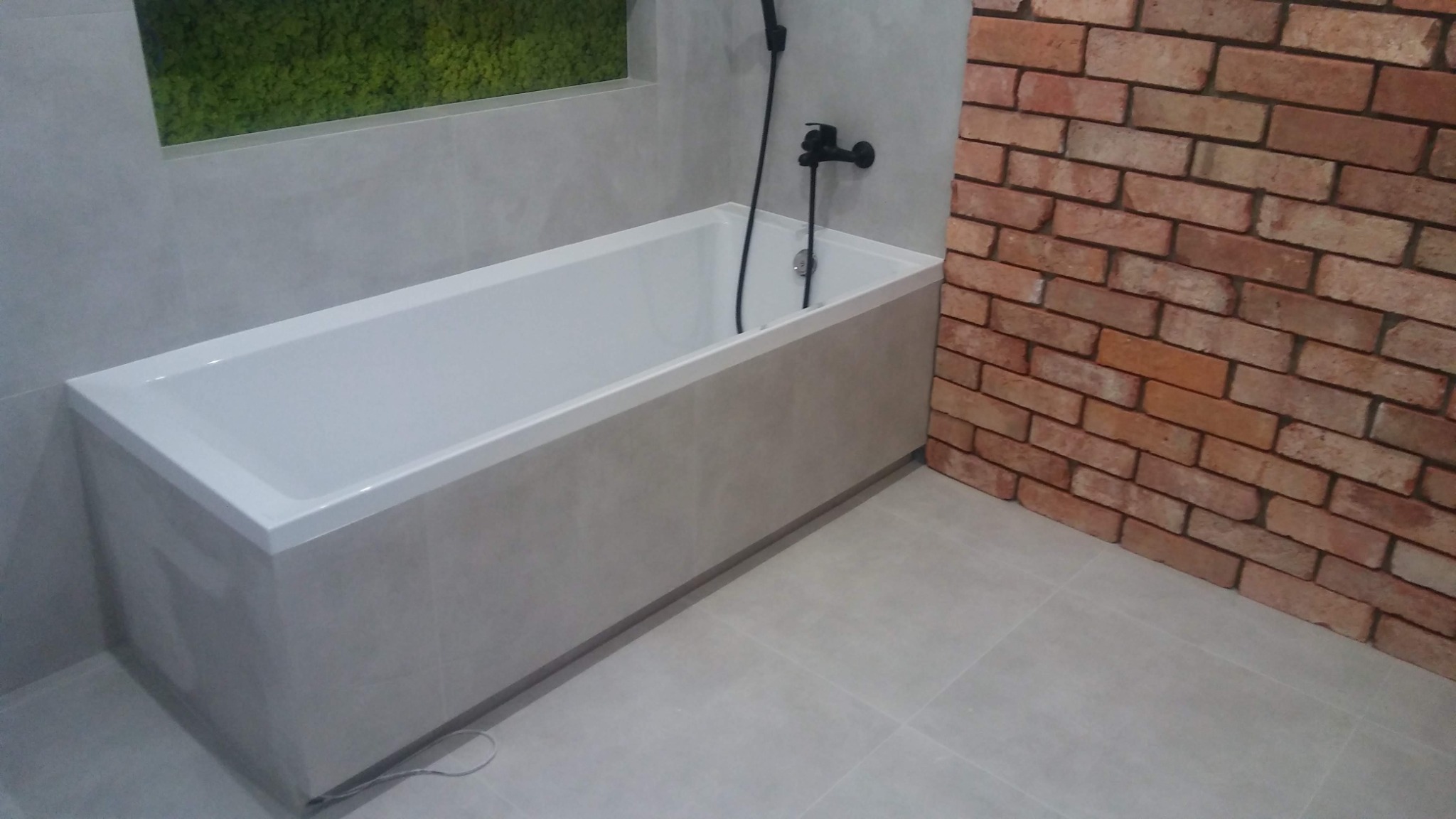 Nowoczesna łazienka z białą wanną obudowaną płytkami w odcieniach szarości, czarną armaturą i ścianą wykończoną imitacją cegły, z zielonym mchem w dekoracyjnej wnęce.