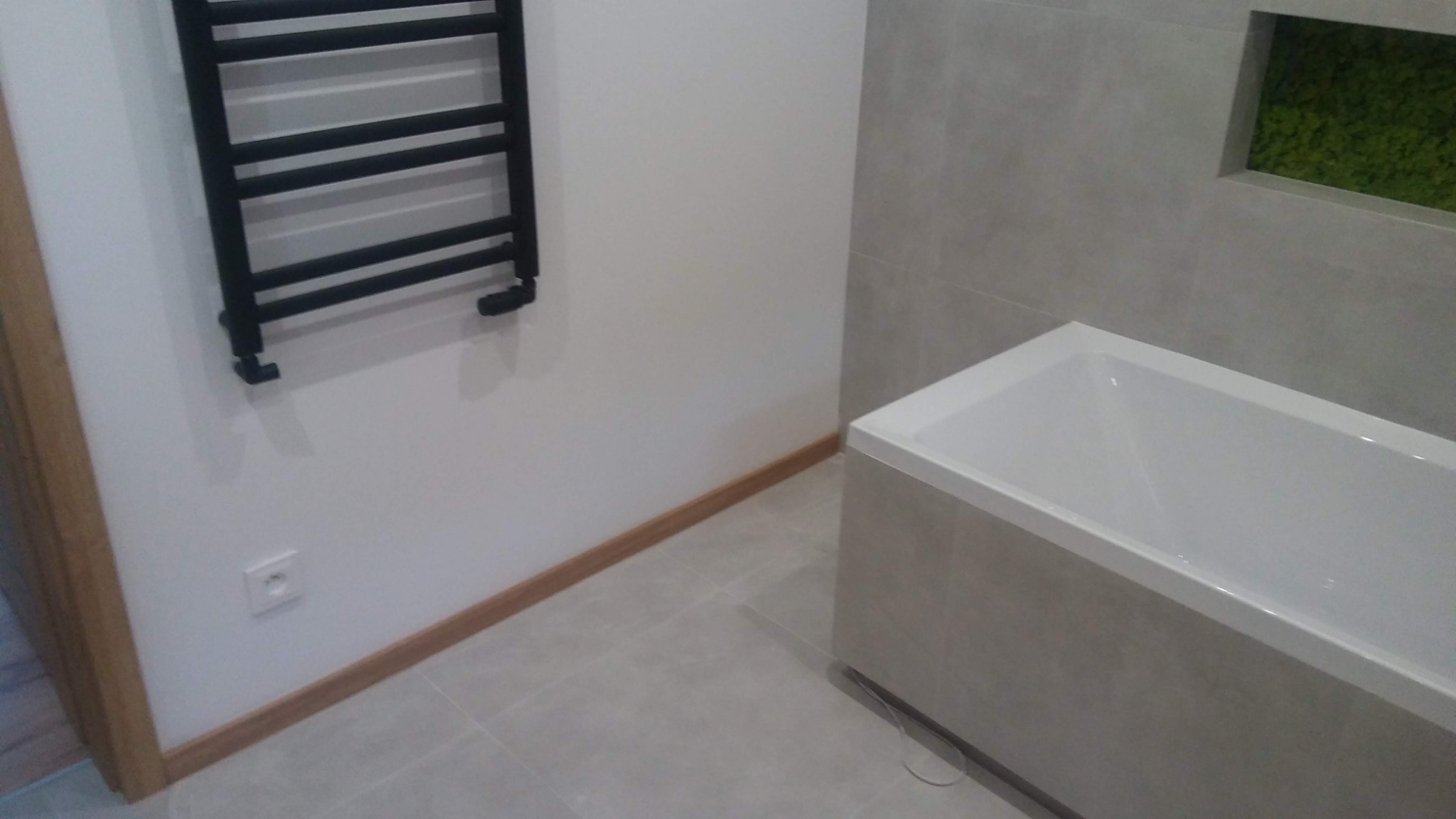 Nowoczesna łazienka z wanną w zabudowie z szarych płytek, czarnym grzejnikiem drabinkowym na białej ścianie, szarą podłogą i dekoracyjną wnęką z zielonym mchem.