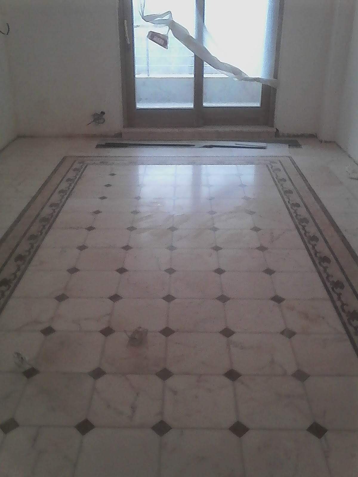 Ułożona marmurowa podłoga z dekoracyjnym bordiurem, widok na okno z rozwianą firanką, surowy stan wykończenia pomieszczenia.