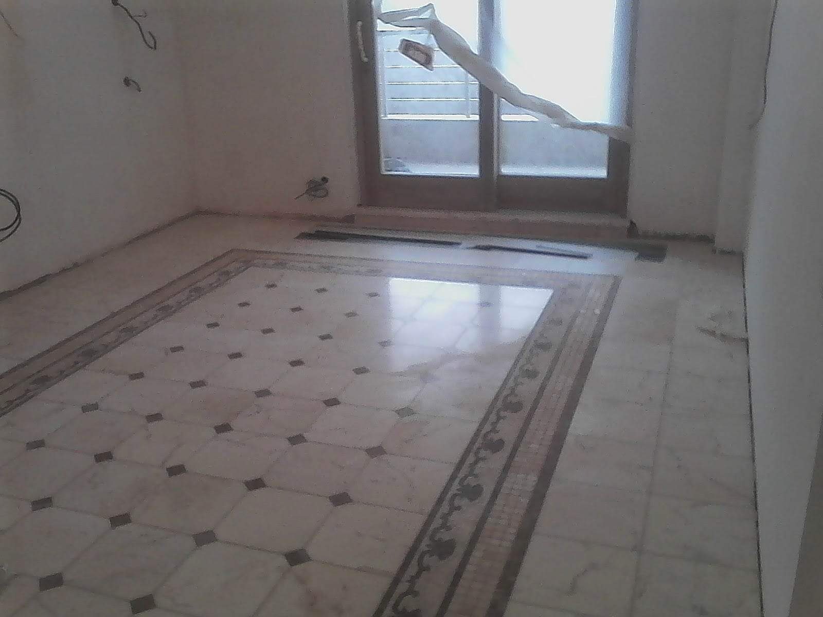 Wykończona podłoga z jasnych płytek z ciemnymi wstawkami i dekoracyjnym obramowaniem, widoczne przygotowania do dalszych prac wykończeniowych, okno zasłonięte folią malarską.