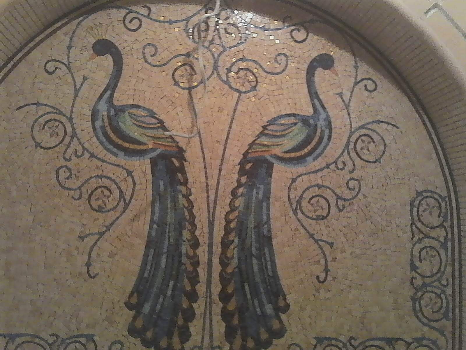 Mozaika ścienna przedstawiająca dwa pawie z piórami w odcieniach niebieskiego, zieleni i pomarańczy na beżowym tle z dekoracyjnymi ornamentami.