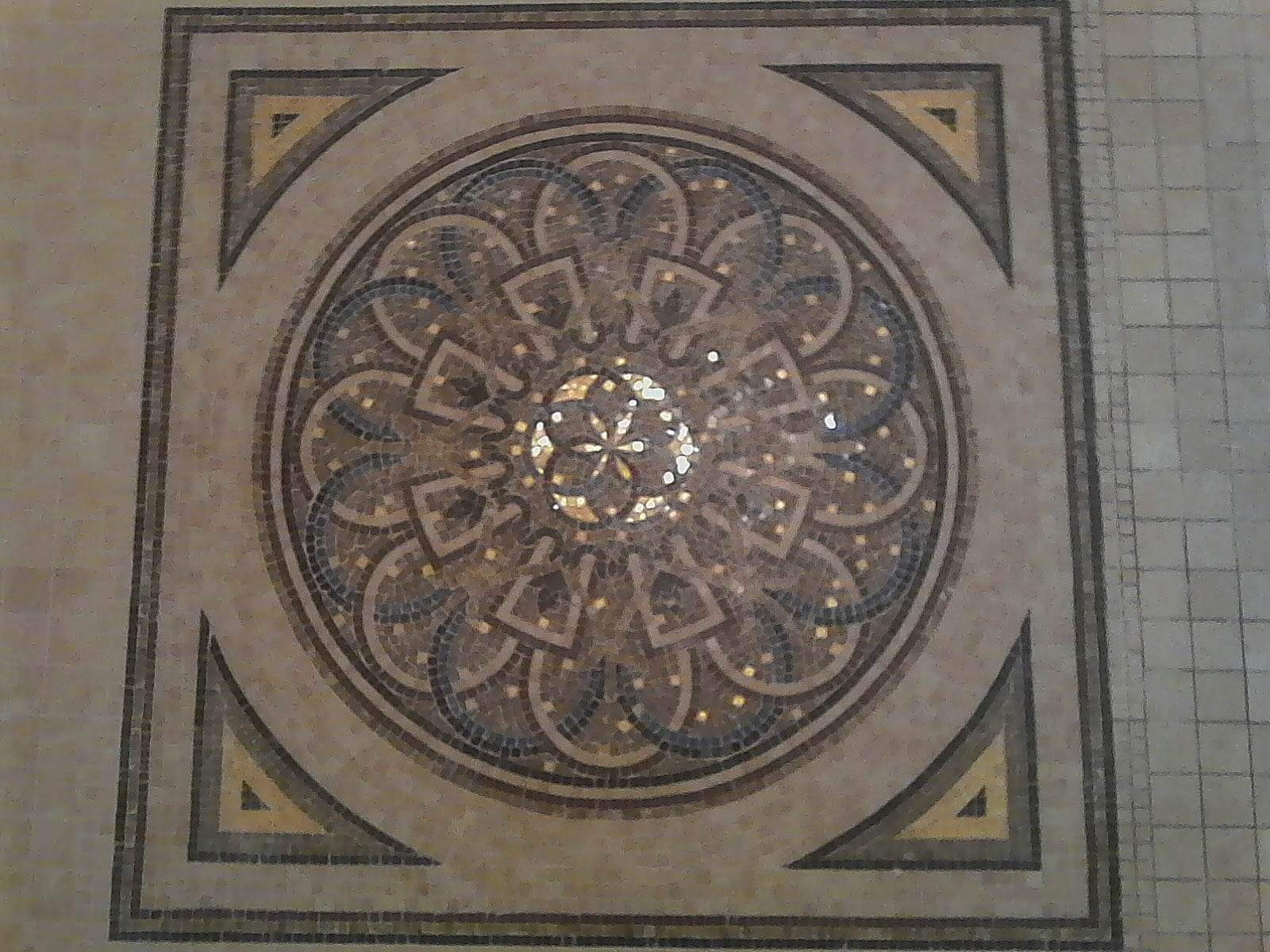 Dekoracyjna mozaika podłogowa w kształcie rozety, z elementami w kolorach beżu, brązu, złota i granatu, umieszczona centralnie w kwadratowej ramie z motywem trójkątów w narożnikach.