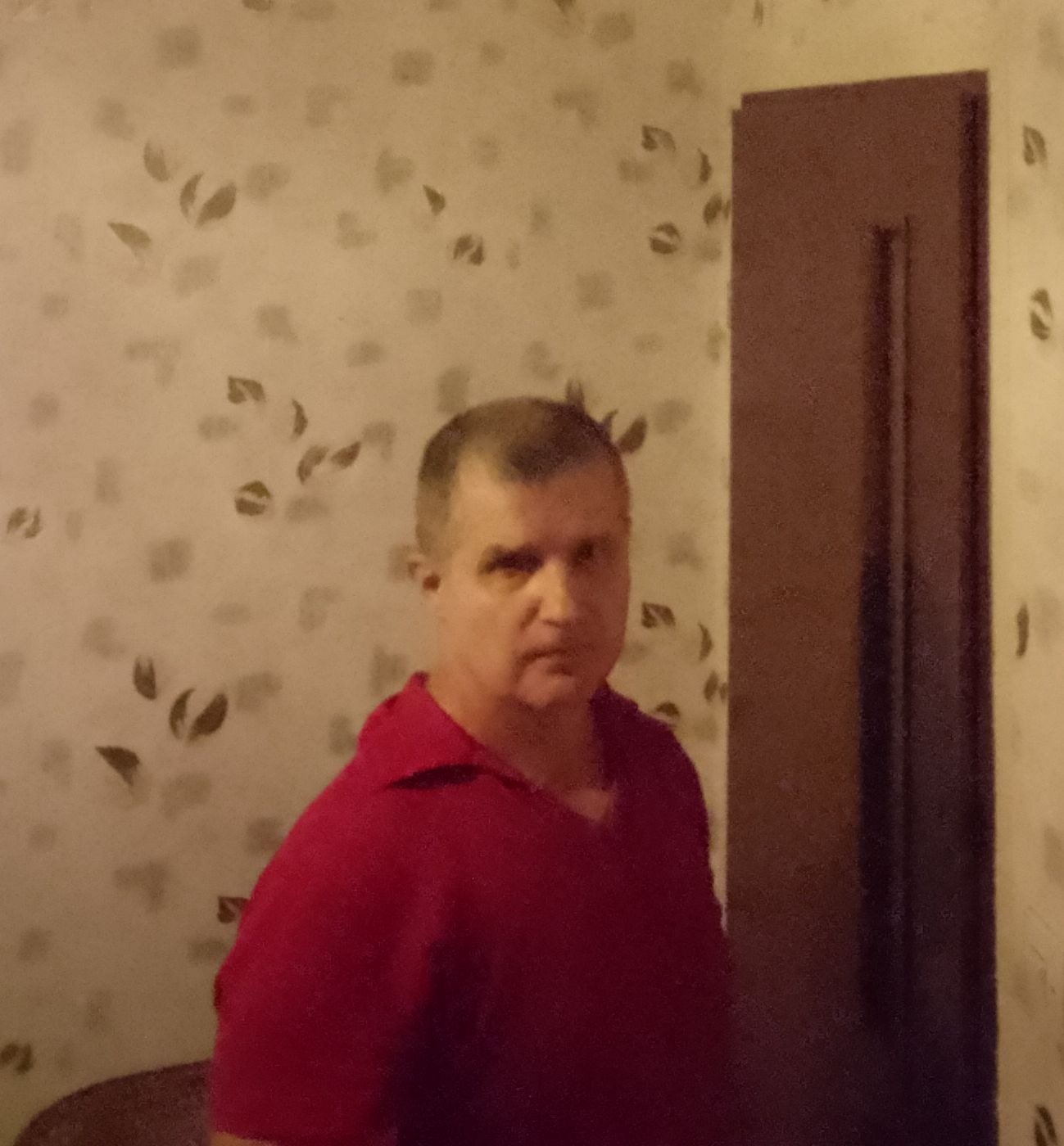 Mężczyzna w czerwonej koszulce polo stoi na tle ściany z tapetą w liście i brązowych drzwi.