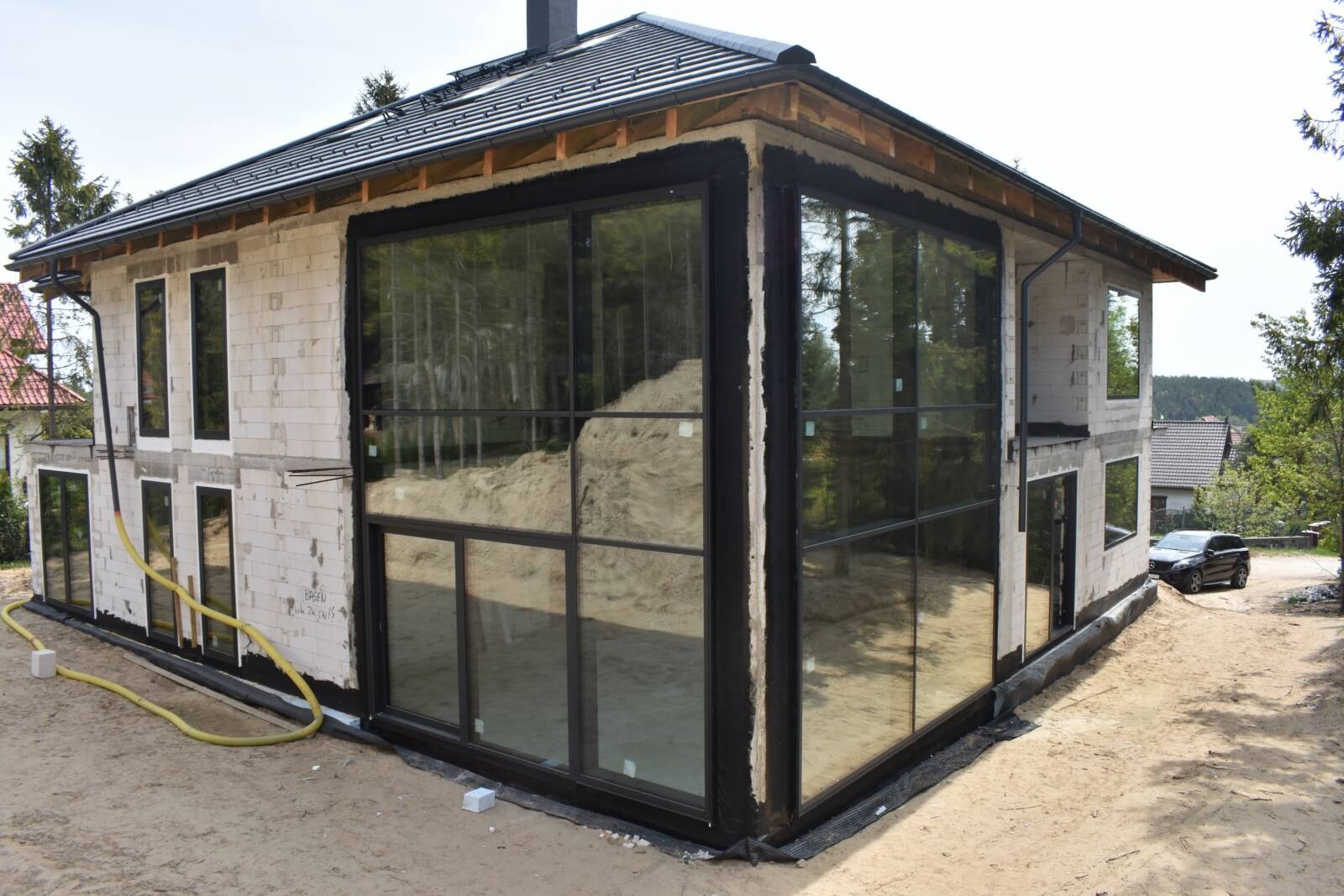 Dom w trakcie budowy z dużymi oknami z czarnymi ramami, widoczny surowy mur z betonu komórkowego i żółty wąż na piasku.