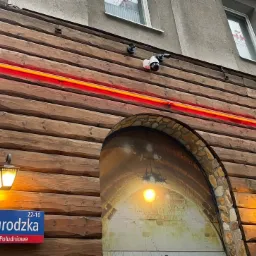 Zewnętrzny montaż trzech kamer monitoringu na drewnianej elewacji budynku z czerwoną neonową linią i dekoracyjnym wejściem imitującym tunel.