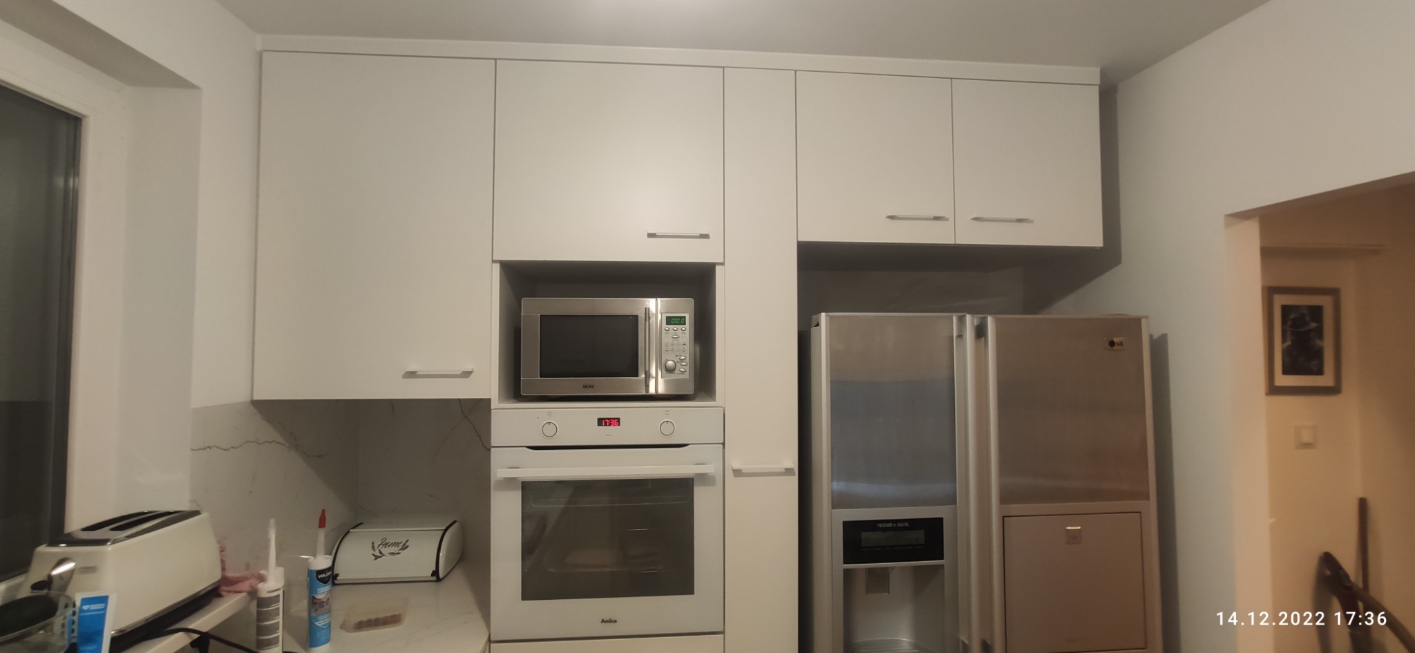 Biała zabudowa kuchenna z szafkami wiszącymi, piekarnikiem, mikrofalówką i lodówką, widok na blat z tosterem i akcesoriami montażowymi, marmurkowy wzór na ścianie.