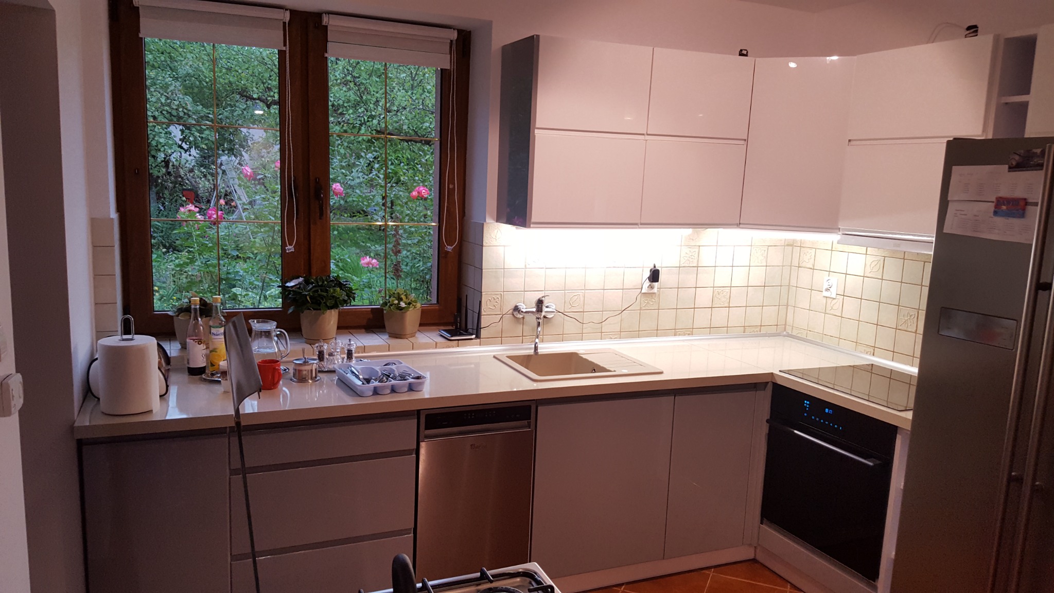 Jasna kuchnia z białymi szafkami i blatem w Koszalinie, z oknem na ogród z kwitnącymi różami, widok z perspektywy blatu z akcesoriami kuchennymi.