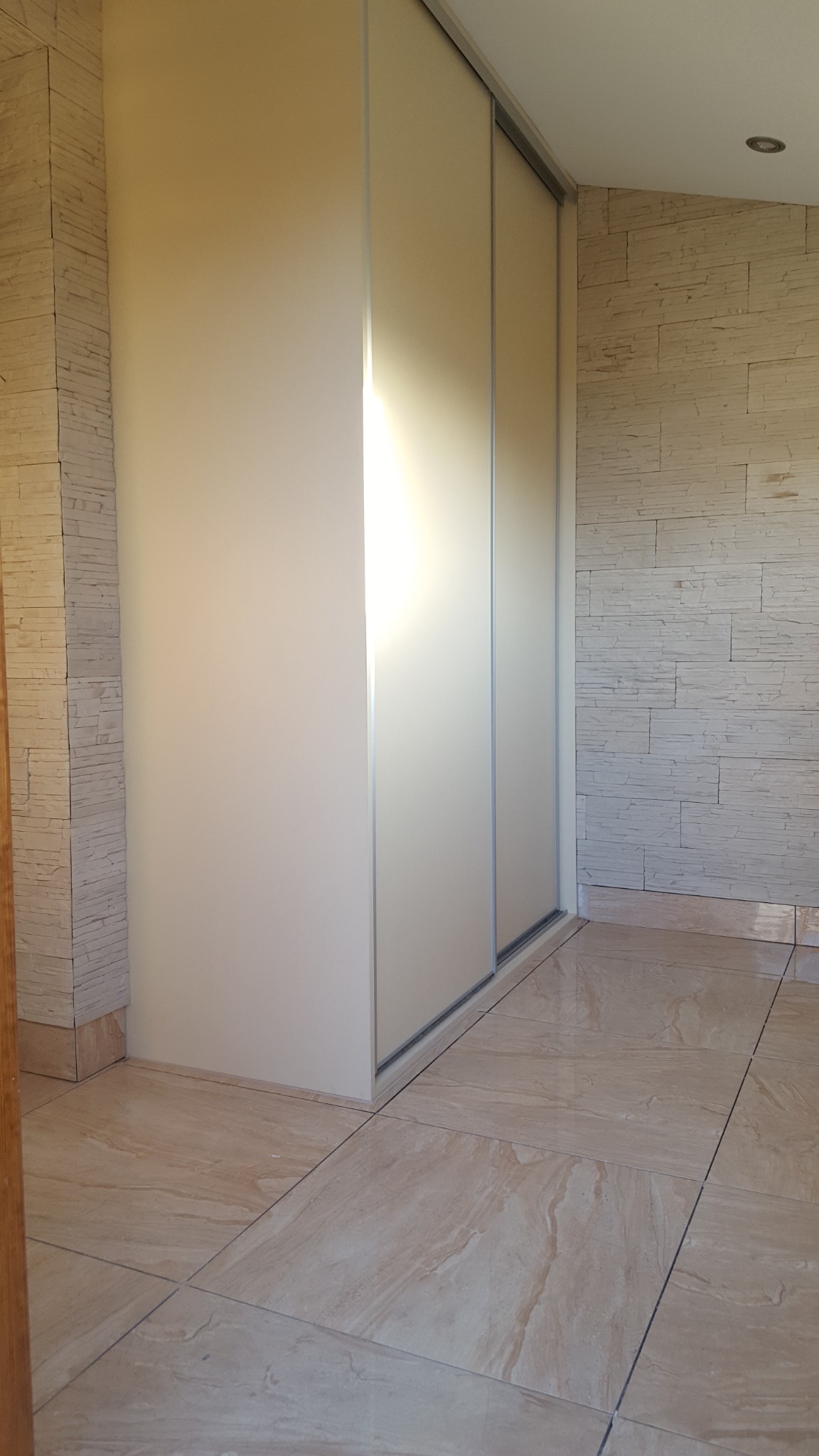 Biała szafa przesuwna z lustrzanymi drzwiami wbudowana we wnękę, wykończenie ściany imitujące kamień, jasna podłoga z płytek.