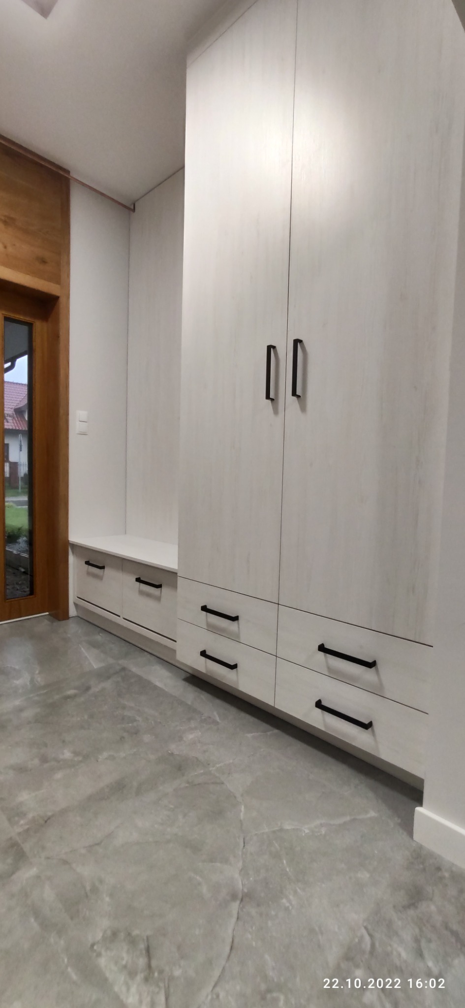 Biała szafa z szufladami i szafkami z czarnymi uchwytami, połączona z niską zabudową z siedziskiem w przedpokoju, widok na szarą podłogę z płytek imitujących marmur.