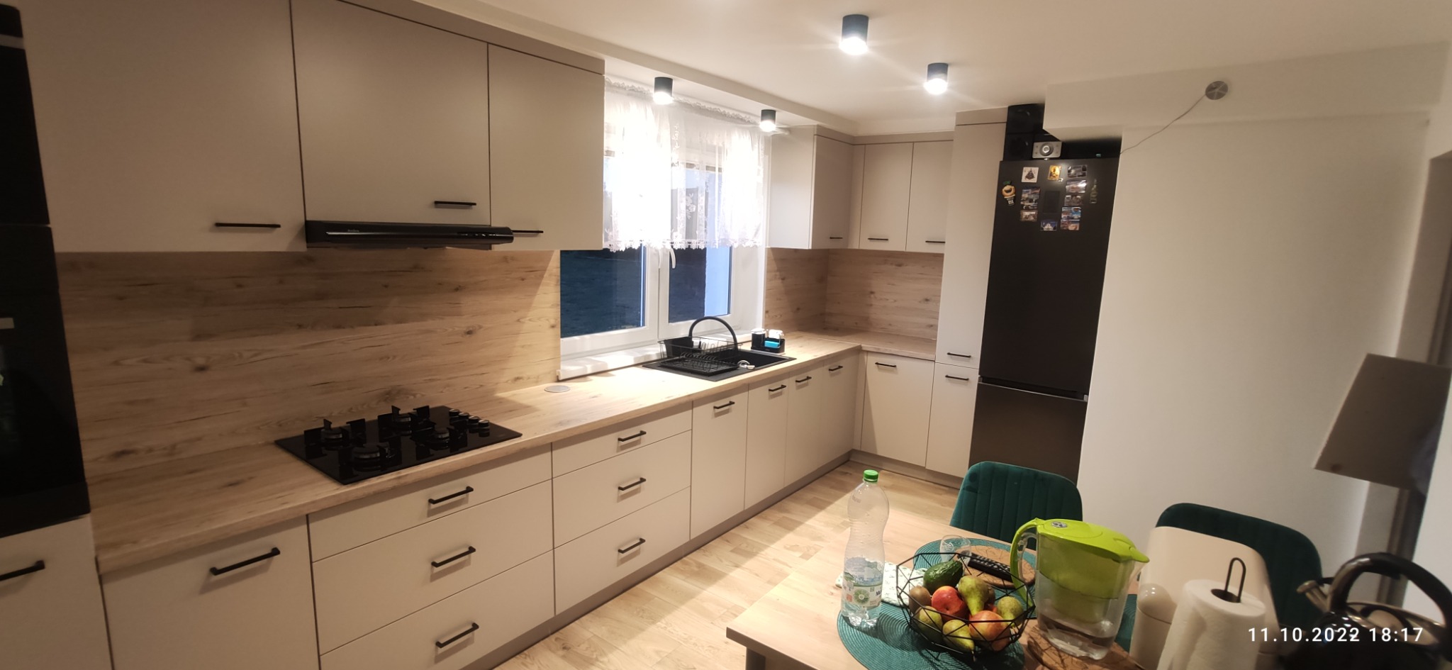 Jasna kuchnia z drewnianym blatem i czarnymi uchwytami, widok na płytę grzewczą, zlew, lodówkę i stół z owocami.