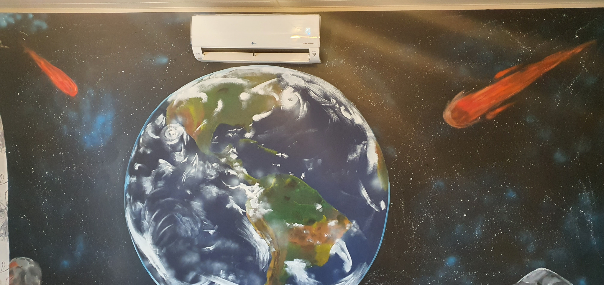 Biała klimatyzacja LG zamontowana na ścianie z ręcznie malowanym motywem kosmicznym, przedstawiającym planetę Ziemię i czerwone komety na tle ciemnej przestrzeni kosmicznej.