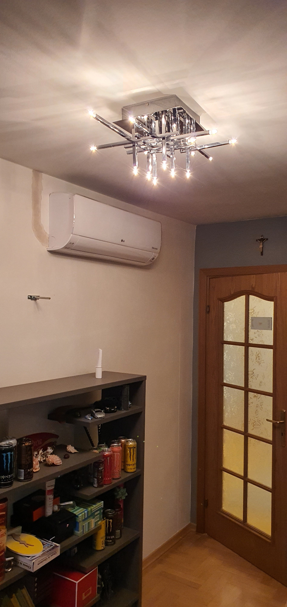 Biała jednostka klimatyzacji LG zamontowana na ścianie w pokoju, pod nią półka z różnymi przedmiotami, nad nią nowoczesna lampa sufitowa, obok drewniane drzwi z szybami.