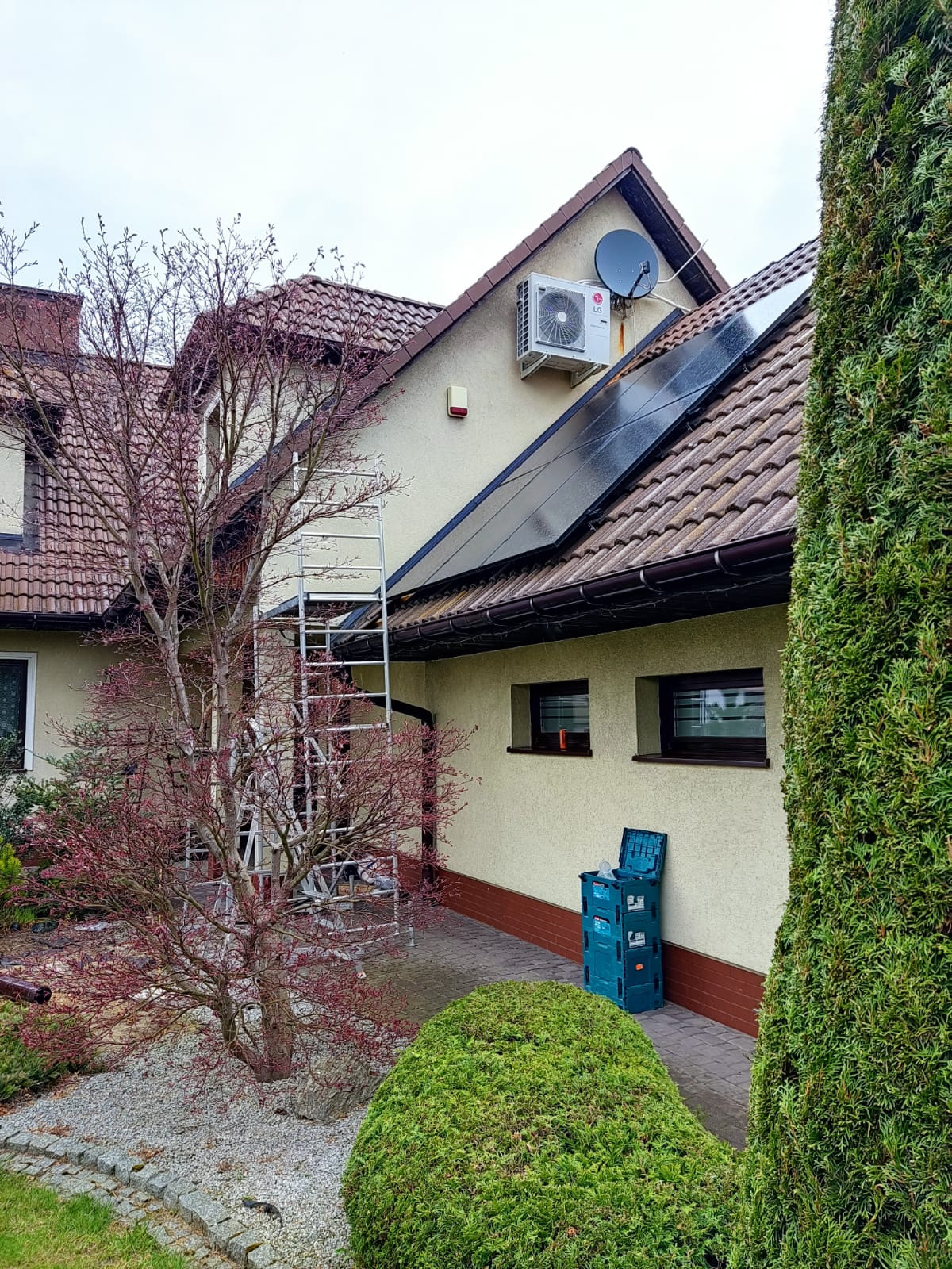 Klimatyzator LG zamontowany na elewacji domu jednorodzinnego, obok panele fotowoltaiczne, drabina aluminiowa oparta o ścianę, skrzynki narzędziowe na chodniku.