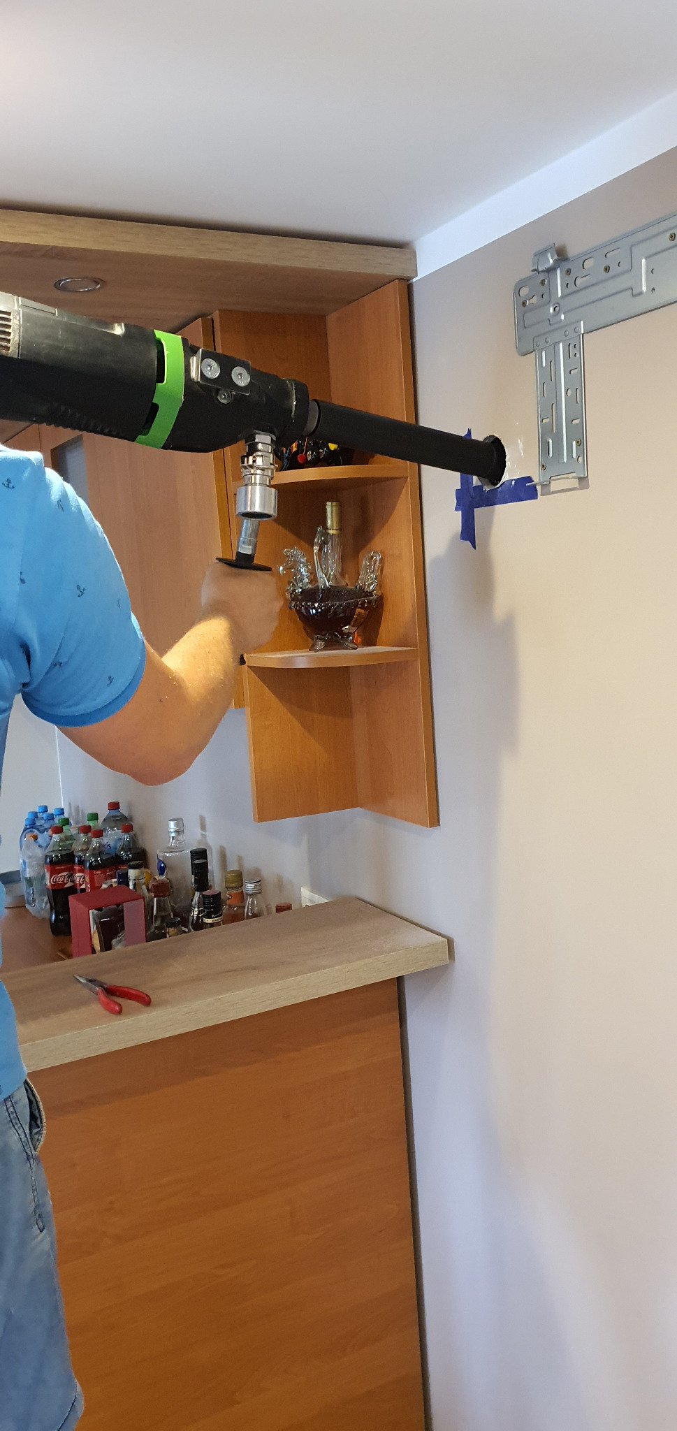 Instalator wierci otwór w ścianie pod rurę klimatyzacji, widoczny uchwyt montażowy klimatyzatora, narzędzia i butelki na blacie.