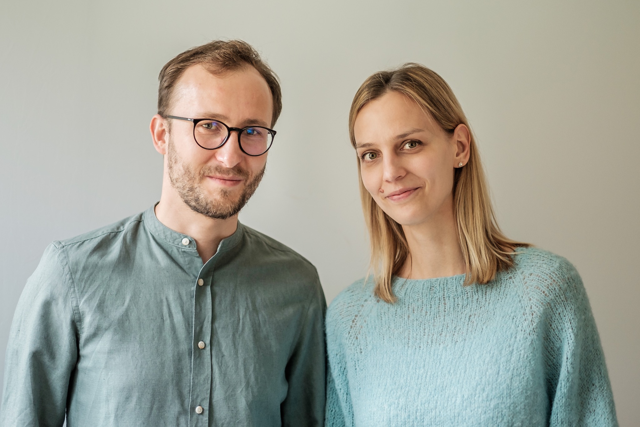 Portret dwojga uśmiechniętych osób: mężczyzny w okularach i lnianej koszuli oraz kobiety w błękitnym swetrze, na neutralnym tle.