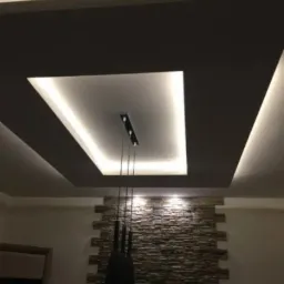 Nowoczesny sufit podwieszany z oświetleniem LED w kształcie kwadratu i wisząca lampa na tle ściany z kamienia dekoracyjnego.