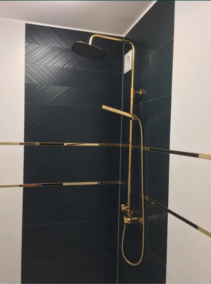 Złota armatura prysznicowa na tle czarno-białych płytek z delikatnym wzorem jodełki, wykończona złotymi listwami dekoracyjnymi.