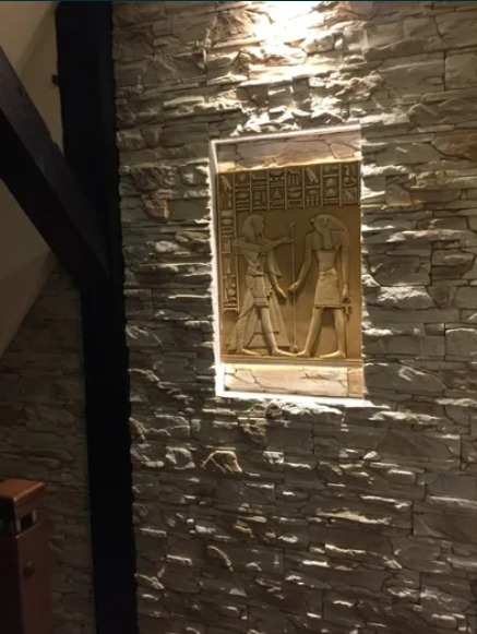 Ściana z kamienia dekoracyjnego z podświetloną niszą zawierającą płaskorzeźbę w stylu egipskim, widoczny fragment drewnianej konstrukcji.