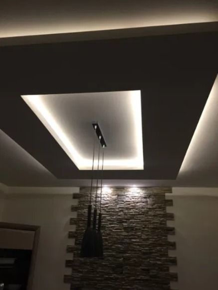 Nowoczesny sufit podwieszany z oświetleniem LED w kształcie kwadratu i wisząca lampa na tle ściany z kamienia dekoracyjnego.