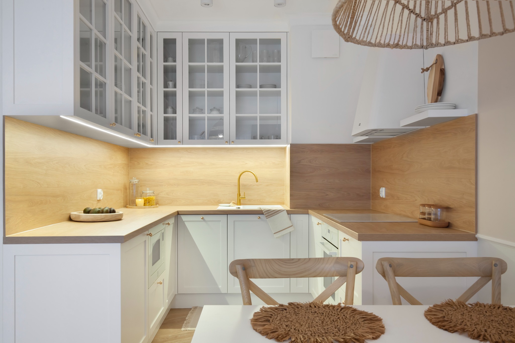 Jasna kuchnia w stylu skandynawskim z białymi szafkami, drewnianymi blatami i złotą armaturą, oświetlona listwami LED pod szafkami wiszącymi. Na blacie drewniana misa z awokado i szklane słoje.