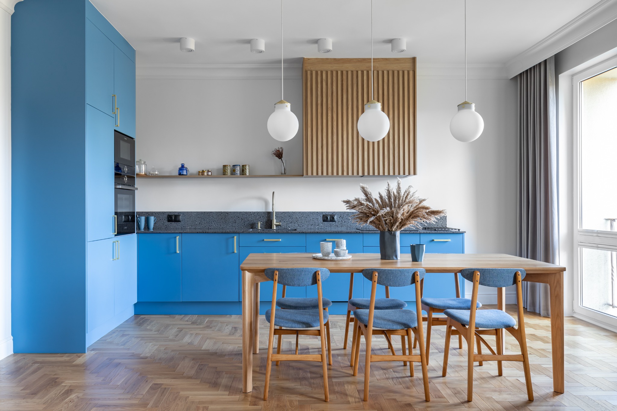 Jasne wnętrze kuchni w Gdańsku z niebieskimi szafkami, drewnianym stołem i krzesłami z niebieską tapicerką oraz parkietem ułożonym w jodełkę.
