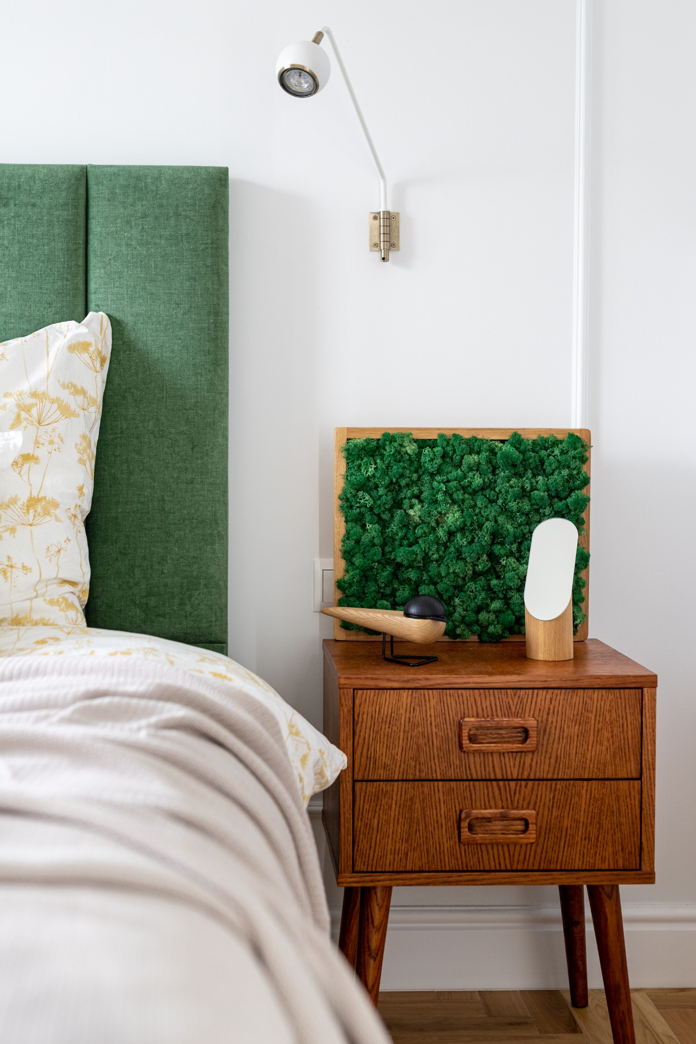 Szafka nocna w stylu mid-century modern z drewnianą lampką i panelem z mchu na tle zielonego zagłówka łóżka i białej ściany z listwami.