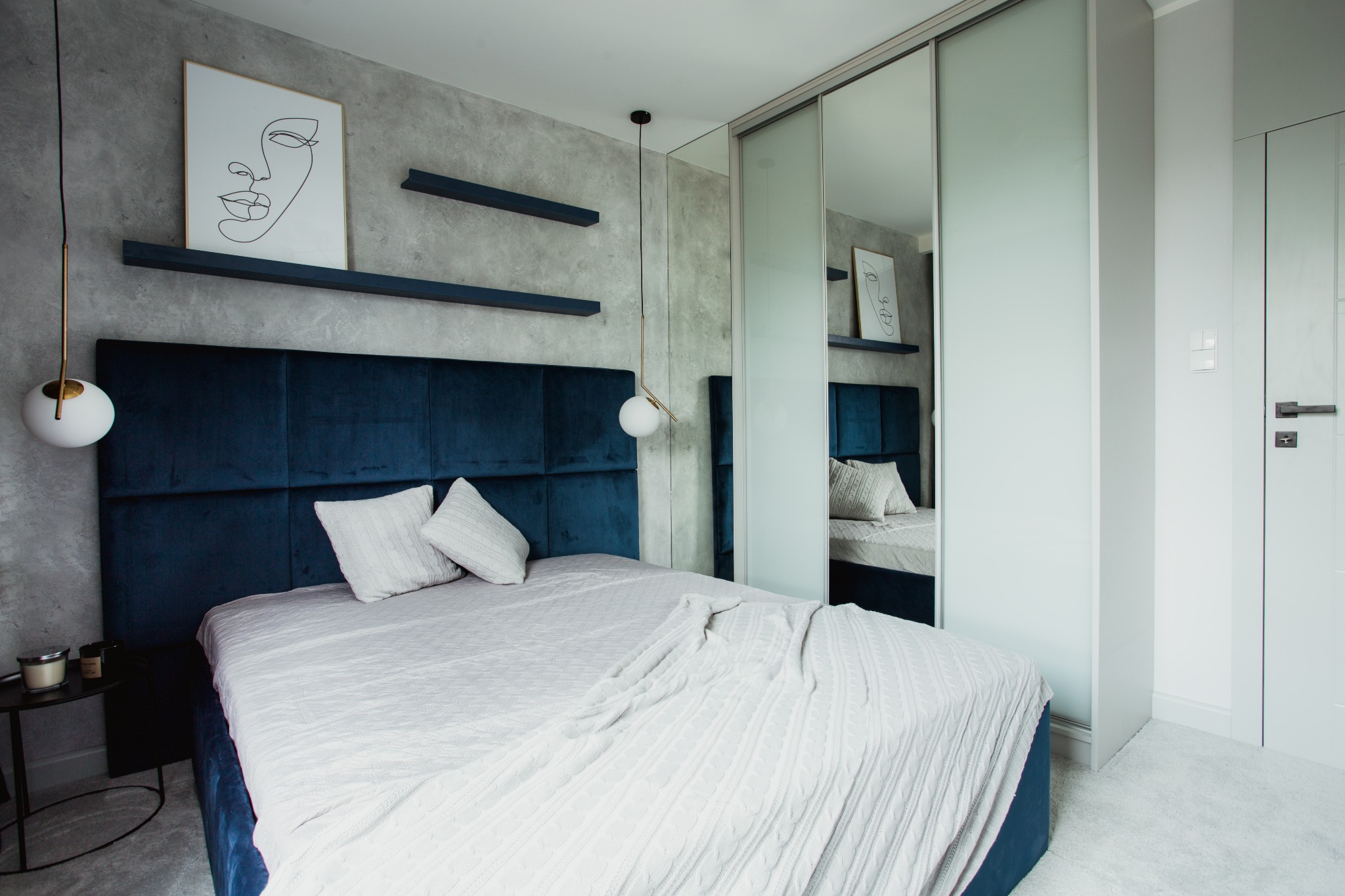 Sypialnia w stylu soft loft z ciemnoniebieskim tapicerowanym zagłówkiem, szarą ścianą z półkami, grafiką liniową i lampą wiszącą z kulistym kloszem, odbicie w lustrzanych drzwiach szafy.