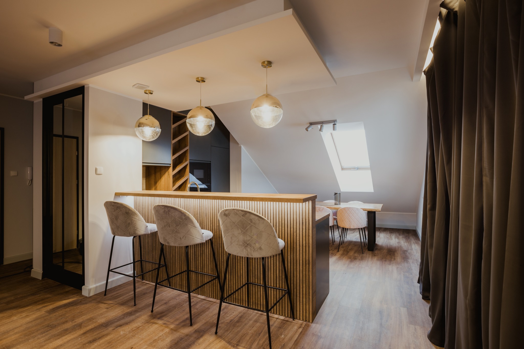 Wnętrze mieszkania w stylu soft loft: wysoki barek z trzema welurowymi hokerami na tle drewnianej podłogi, z widokiem na stół jadalny pod oknem dachowym i szklane lampy wiszące.