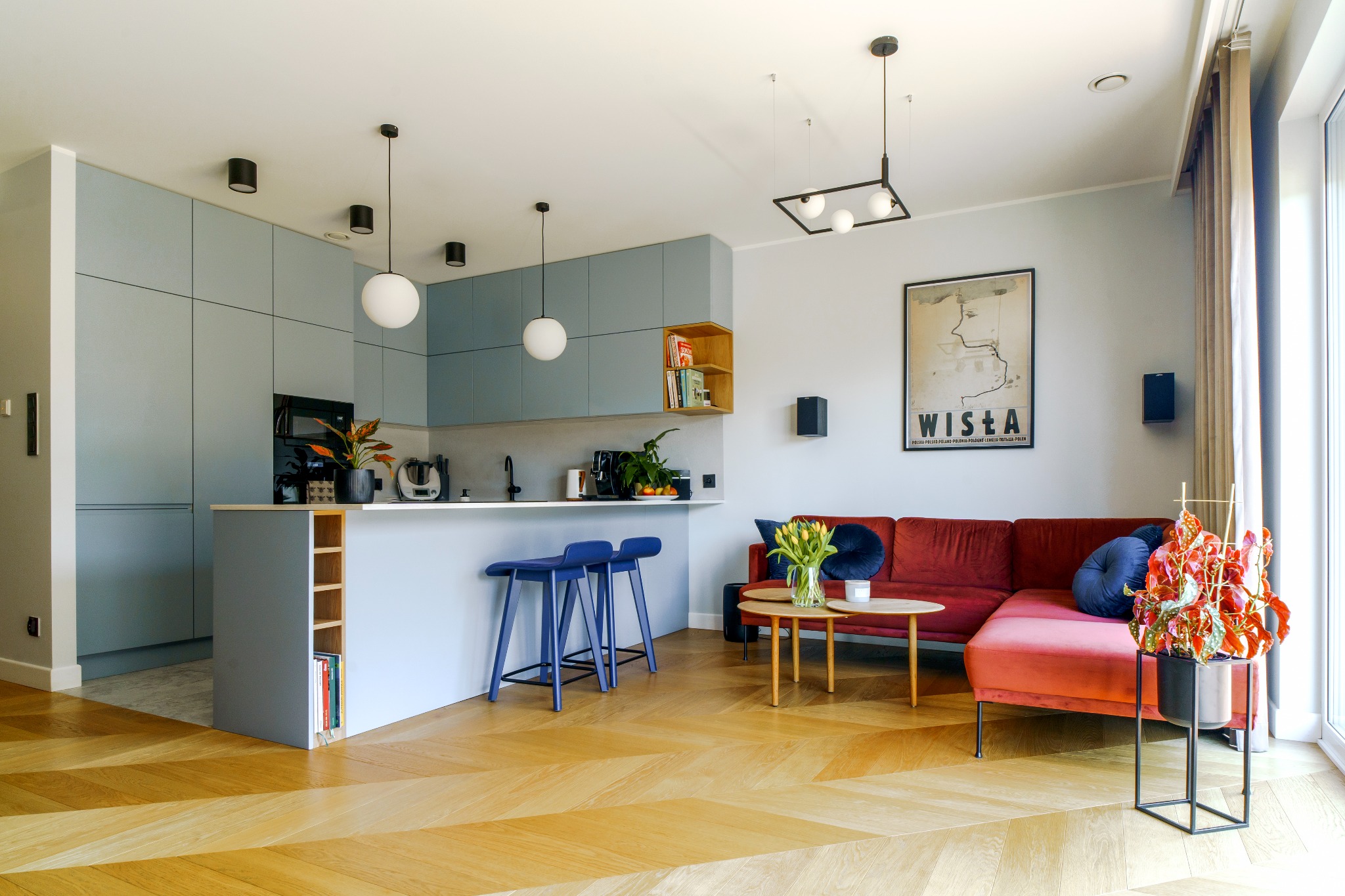 Jasne wnętrze z parkietem w jodełkę, błękitną zabudową kuchenną, wyspą z hokerami, czerwonym narożnikiem i plakatem 'Wisła' na ścianie.