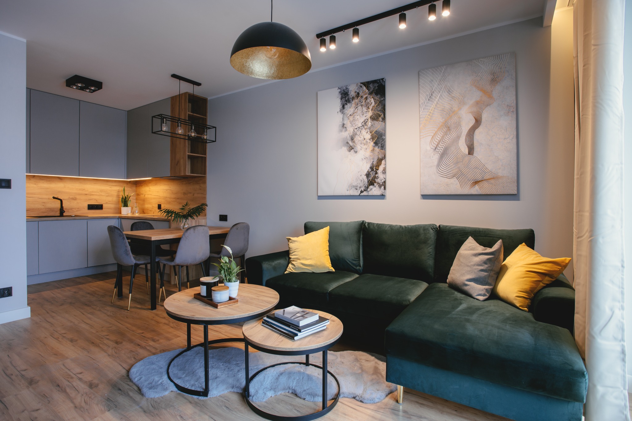 Nowoczesne wnętrze mieszkania w stylu soft loft: zielona sofa narożna z żółtymi poduszkami, okrągłe stoliki kawowe na dywaniku z imitacji futra, drewniany stół z szarymi krzesłami, szara zabudowa...