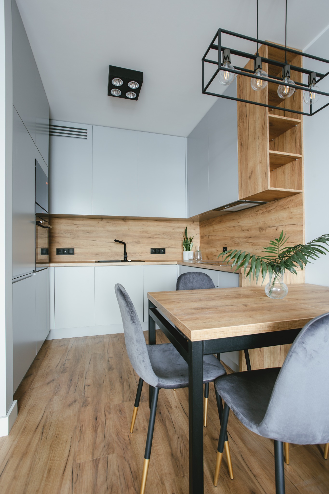 Nowoczesna kuchnia z drewnianymi akcentami: jasnoszare szafki, drewniany panel nad blatem, stół z szarymi krzesłami na złotych nóżkach, oświetlenie sufitowe w stylu loft.