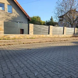 Nowoczesne ogrodzenie z poziomych, szarych paneli między słupkami z piaskowca, widoczne z poziomu kostki brukowej na tle domu i drzew