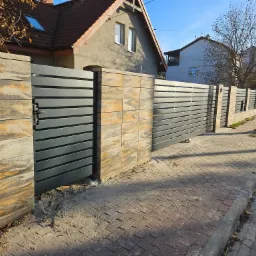 Nowoczesna brama wjazdowa i przęsła ogrodzeniowe z poziomych, szarych profili, osadzone na słupkach z bloczków imitujących piaskowiec, na tle brukowanej ulicy i domów.