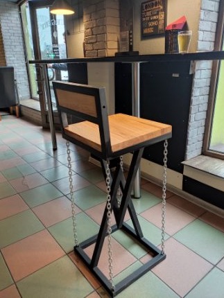Unikalny hoker barowy z drewnianym siedziskiem i oparciem, metalową ramą w kształcie litery X oraz łańcuchami zamiast nóg, ustawiony przy barze w lokalu.