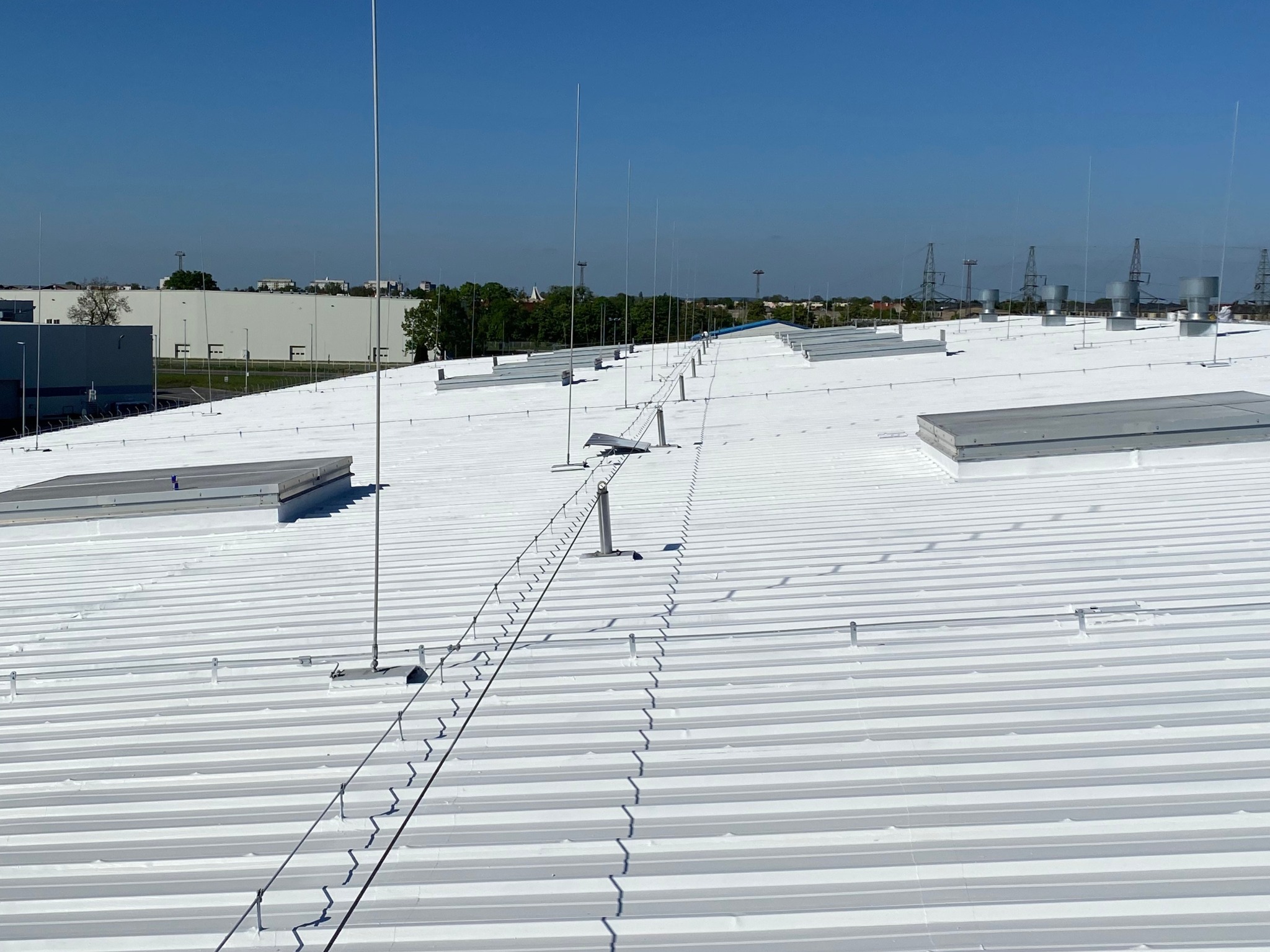 Biały, płaski dach przemysłowy z systemem odgromowym i świetlikami, widziany z góry w słoneczny dzień. W tle budynki i słupy energetyczne.