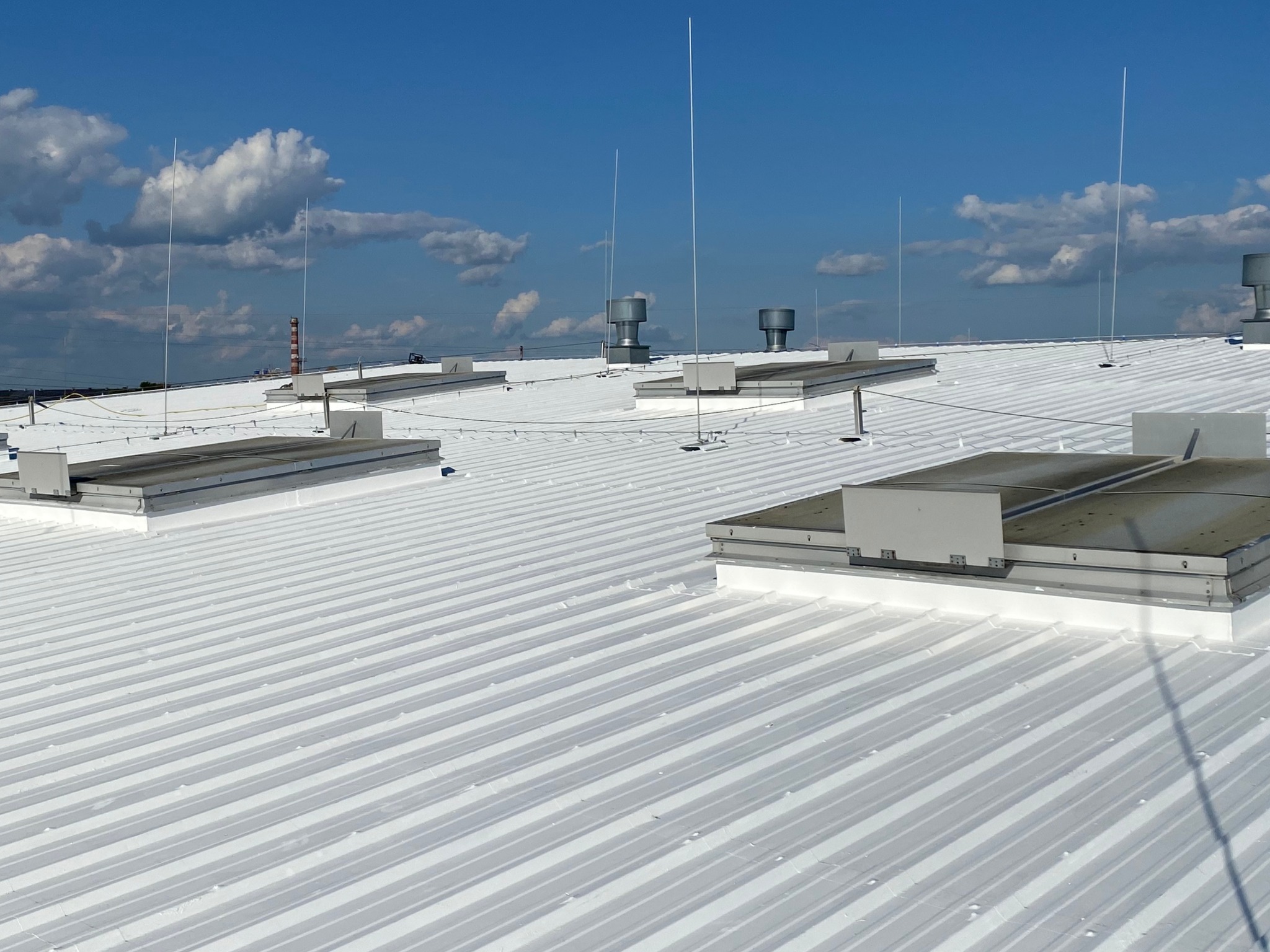 Biały, blaszany dach przemysłowy z odgromnikami i wywietrznikami na tle błękitnego nieba w Zabrzu. Widok z góry, ostre światło słoneczne.