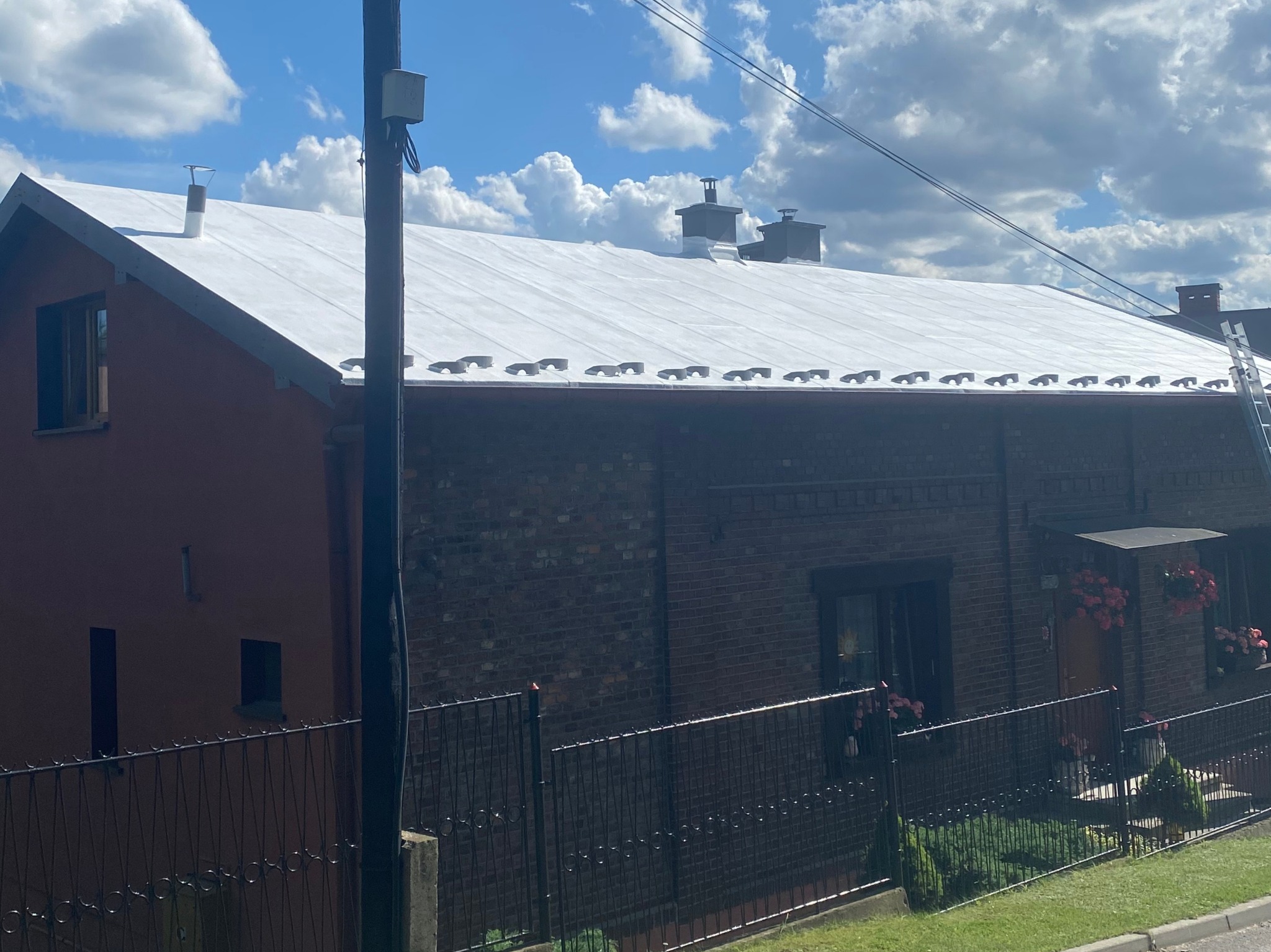 Biały, płaski dach po renowacji na dwukondygnacyjnym budynku z cegły i tynku, widoczny zza czarnego metalowego ogrodzenia w słoneczny dzień.