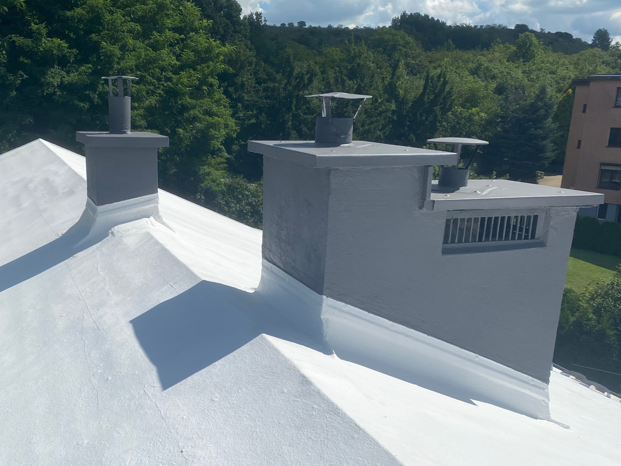 Biały dach po malowaniu z trzema szarymi kominami na tle zieleni. Widoczne detale obróbek blacharskich i wentylacji komina. Jasne, naturalne oświetlenie.