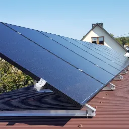 Rząd paneli słonecznych na brązowym dachu z blachy trapezowej, widoczne elementy montażowe i okablowanie, w tle zabudowa jednorodzinna i błękitne niebo.