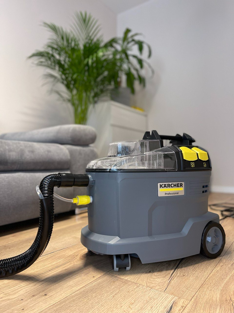 Profesjonalny odkurzacz piorący Kärcher na drewnianej podłodze, z widocznym wężem i żółtą złączką, w tle szara sofa i roślina doniczkowa.