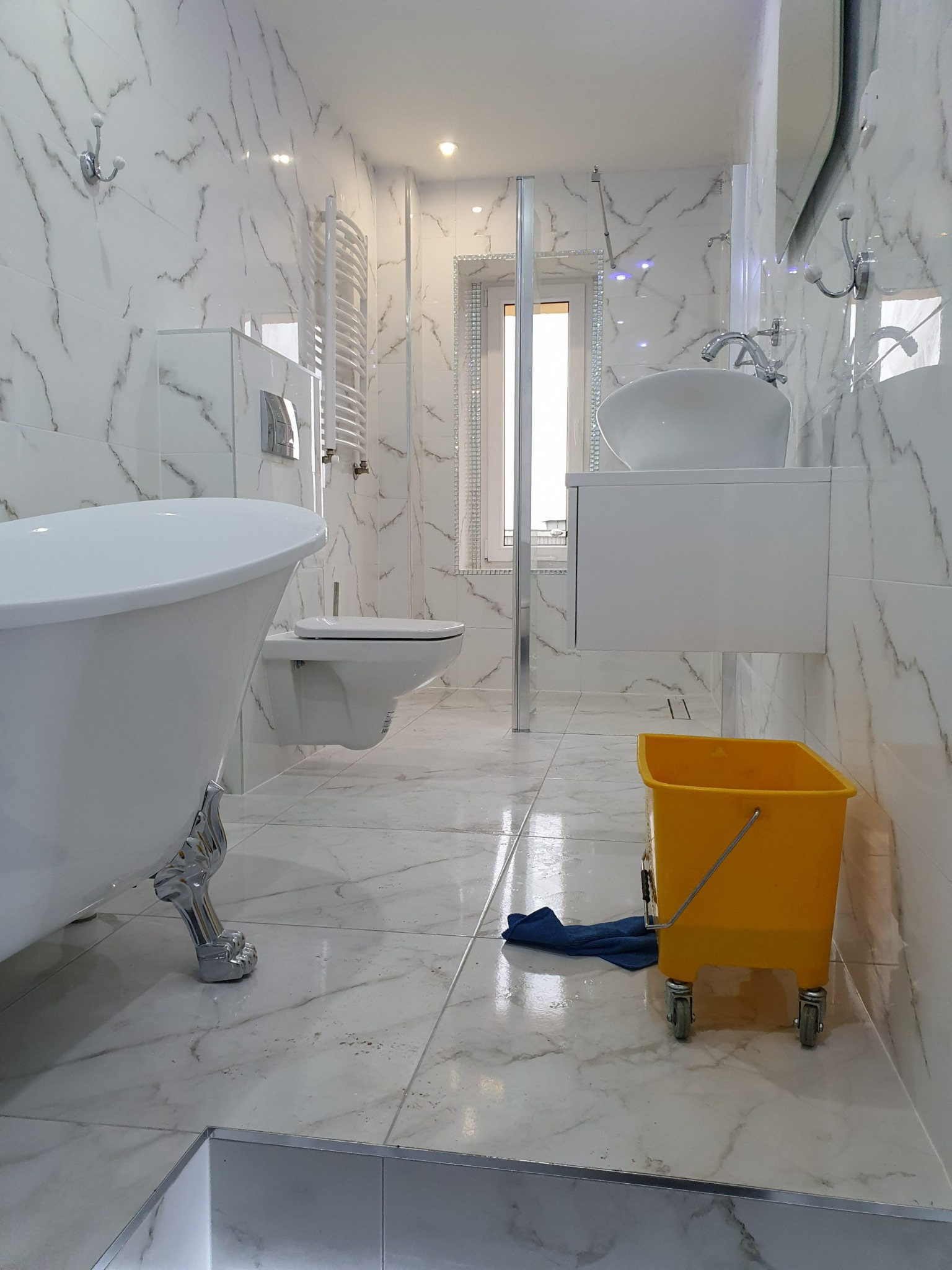 Wnętrze łazienki z białą armaturą, widoczny żółty wózek z mopem i niebieską szmatką na podłodze z marmurowymi płytkami.