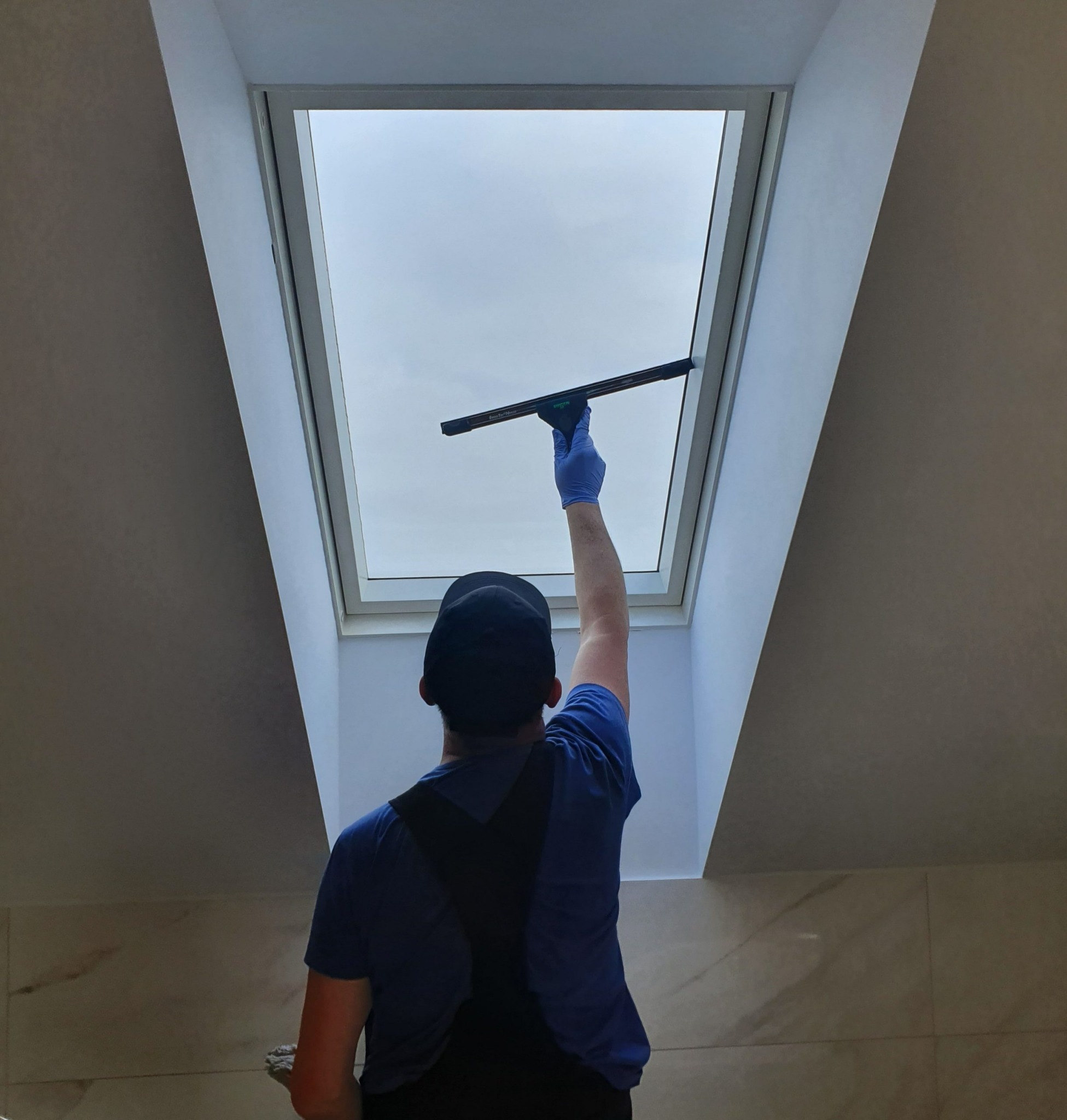 Pracownik w niebieskiej koszulce i czapce, z tyłu, myje okno dachowe za pomocą ściągaczki w niebieskiej rękawicy.