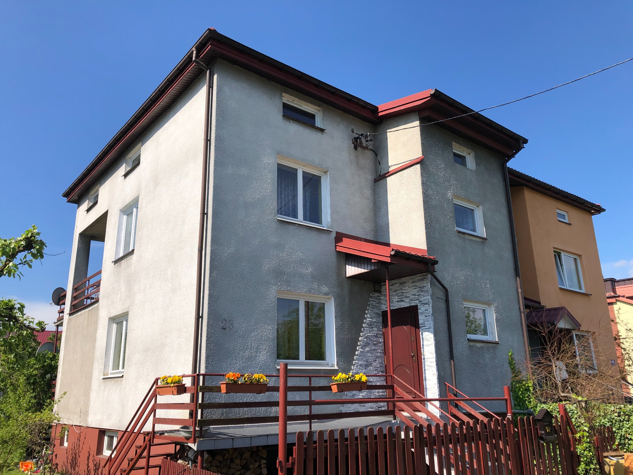 Dwupiętrowy dom z szarą elewacją o nierównej fakturze, czerwonymi elementami wykończeniowymi dachu i balustrad, z drewnem ułożonym pod schodami i kwiatami w doniczkach na balkonie