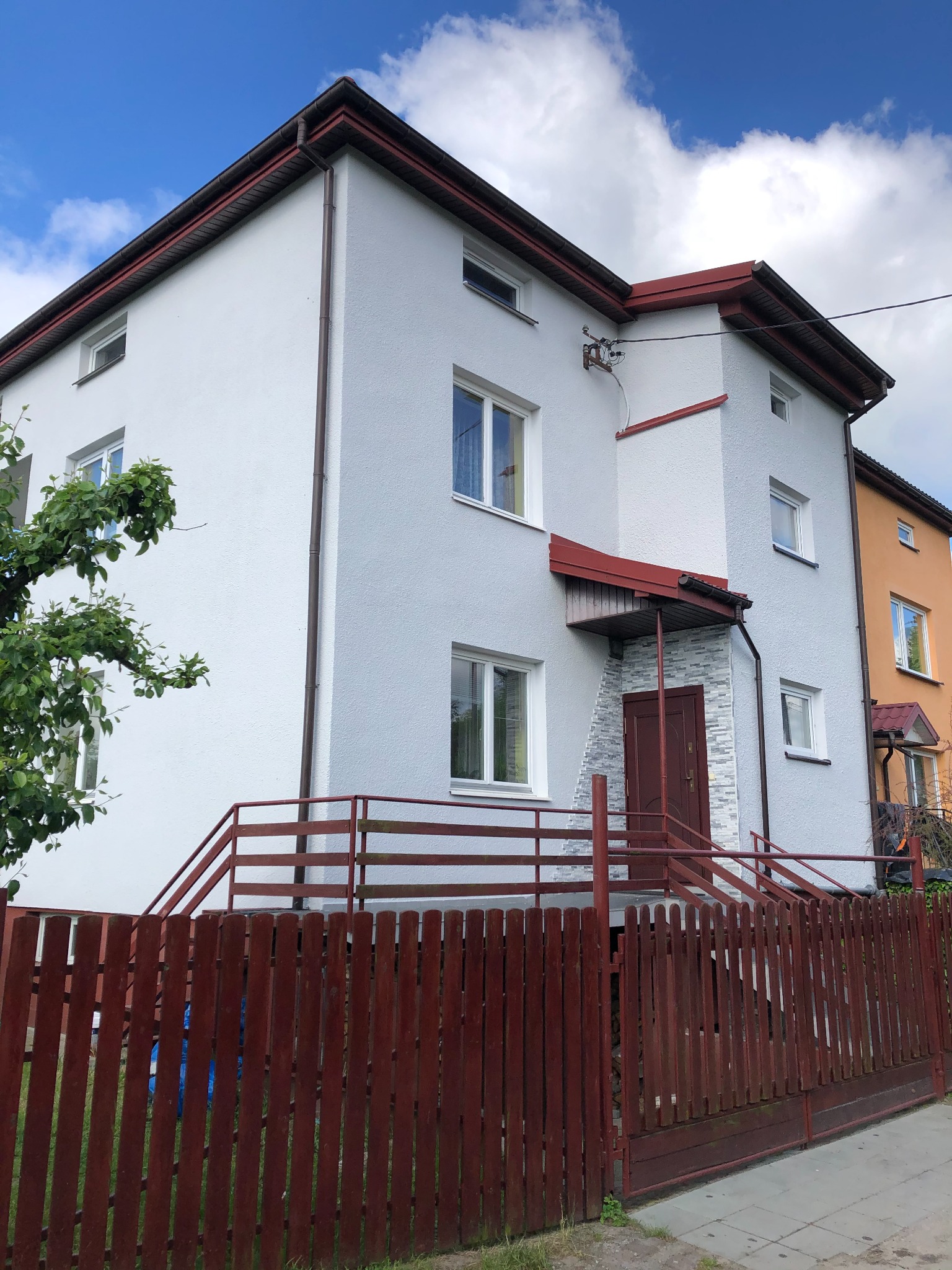 Dwupiętrowy dom z białą elewacją i ciemnoczerwoną stolarką okienną oraz balustradą schodów wejściowych, widziany z poziomu ulicy za drewnianym płotem.