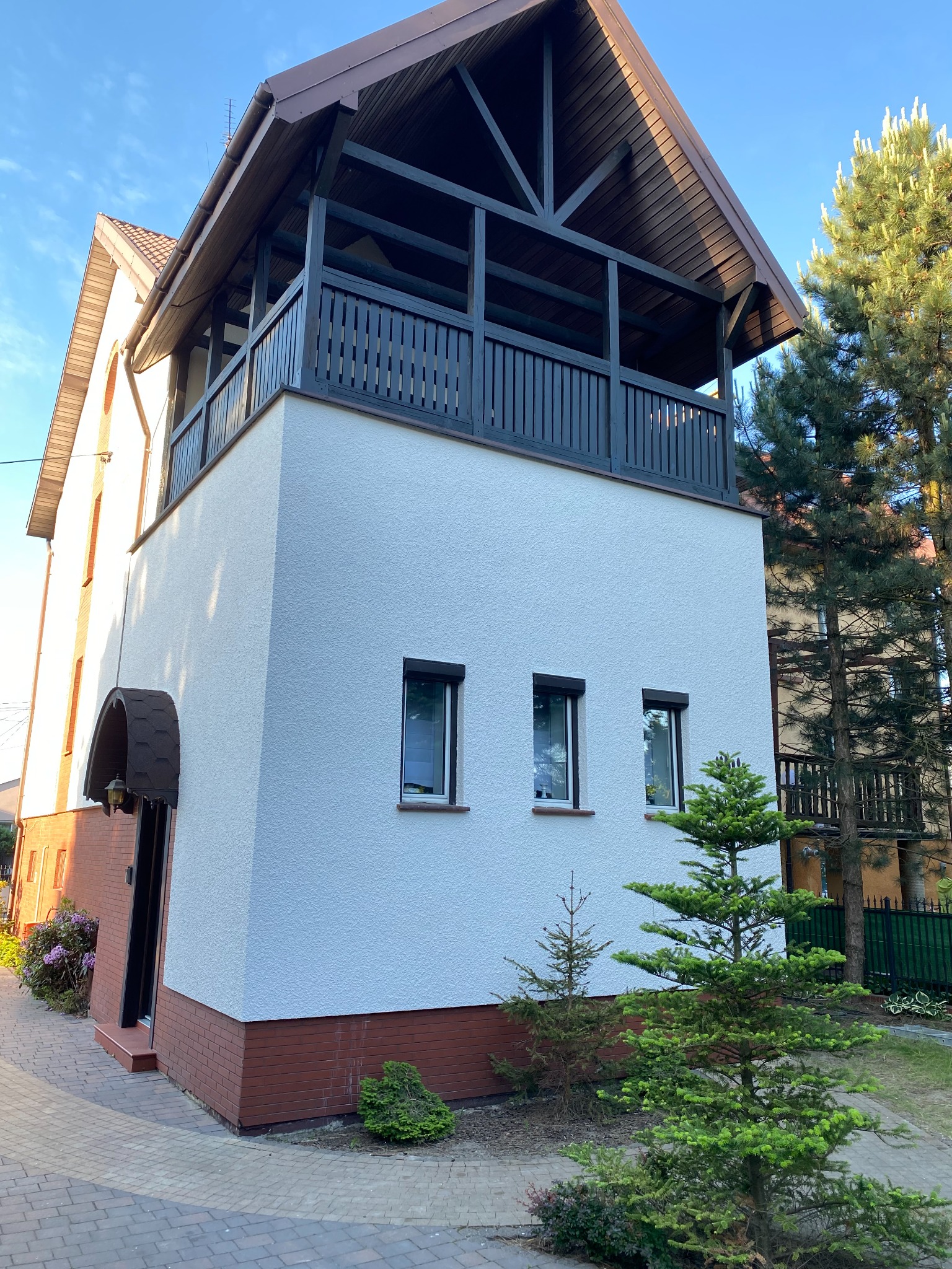 Dwupiętrowy dom z białymi ścianami i ceglaną podstawą, zadaszony balkon z ciemnymi balustradami, widok z poziomu chodnika z kostki brukowej, małe drzewka iglaste w ogródku.