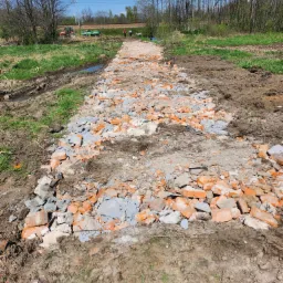 Ścieżka z gruzu ceglanego i betonowego, wstępny etap budowy drogi na podmokłym terenie, w tle traktor z przyczepą i dwie osoby, widoczne ślady opon w błocie.