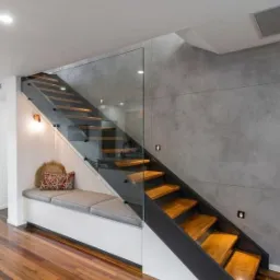 Nowoczesne schody z drewnianymi stopniami, szklaną balustradą i metalową konstrukcją, z siedziskiem pod schodami i oświetleniem punktowym na szarej ścianie.