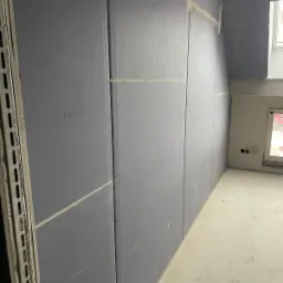 Montaż płyt karton-gips na ścianie pod skosem, widoczne łączenia płyt zaszpachlowane, fragment okna w tle, napis ołówkiem '34/35' na płycie.